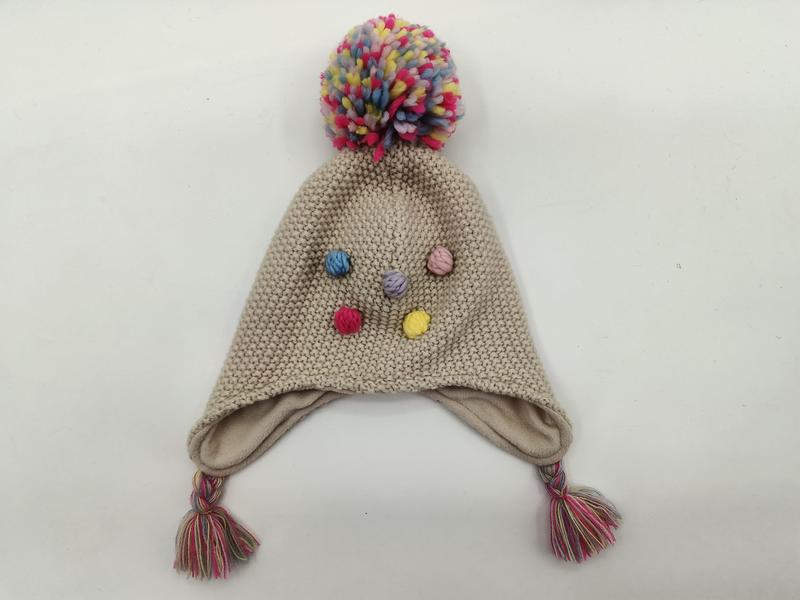 Beige strikket vinterlue til barn med fargerik pom-pom og dusker på sidene.