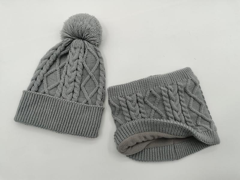 Grå kabelvevd lue med pompom og matchende fleecefôret nakkevarmersett på hvit bakgrunn.