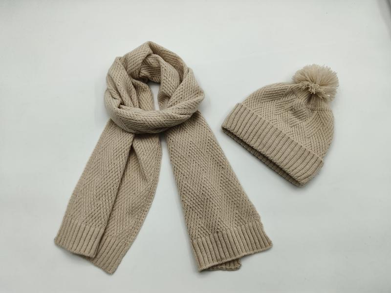 Beige grovstrikket skjerf og matchende lue med pompom på hvit bakgrunn.