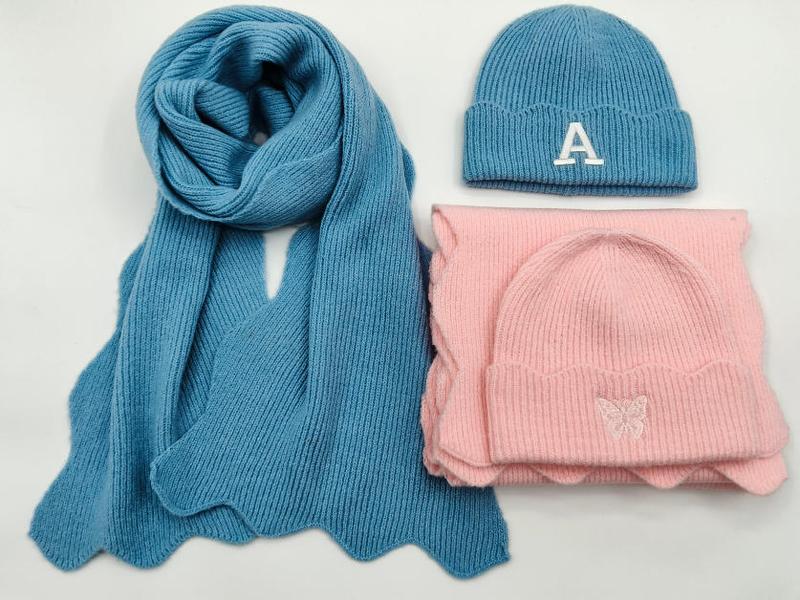 Blå og rosa strikket lue og skjerf-sett med broderte motiver på hvit bakgrunn.