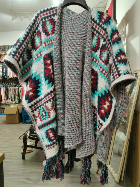 Grå strikket poncho med aztekisk geometrisk mønster i teal, burgunder og hvit, med dusker langs kanten.