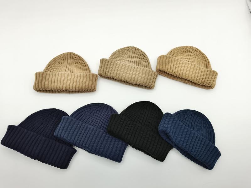 Syv ribbede strikkeluer med oppbrett i beige, marineblå, svart og blå, arrangert på hvit bakgrunn.